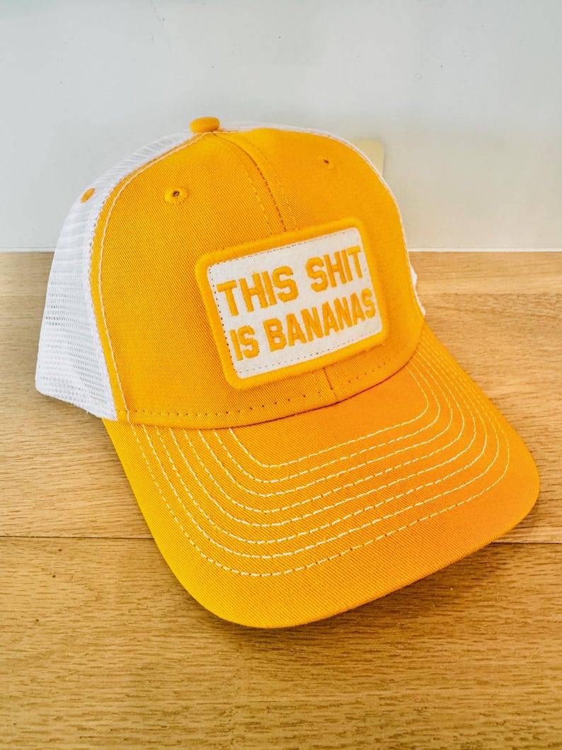 Puede incluir: Gorra de b&eacute;isbol amarilla con la parte trasera de malla blanca. La gorra presenta el texto "THIS SHIT IS BANANAS" en un parche rectangular. Las costuras blancas acent&uacute;an la visera y la parte delantera de la gorra.