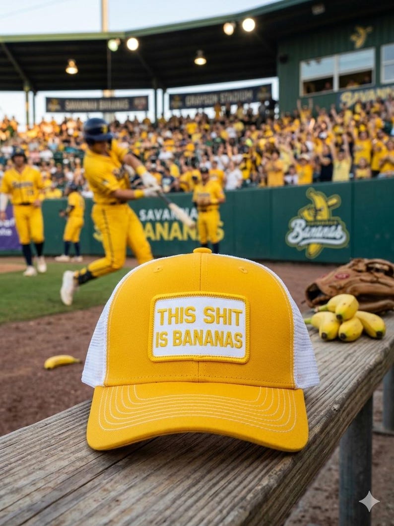 May include: Yellow and white baseball cap with the text "THIS SHIT IS BANANAS" on a patch. The cap is on a wooden bench with a baseball game in the background. A baseball glove and bananas are also visible.