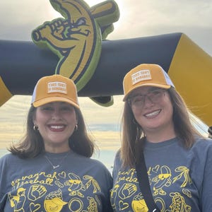 Puede incluir: Dos mujeres con gorras de camionero amarillas y blancas con el texto "THIS SHIT IS BANANAS" y camisetas grises con gr&aacute;ficos de b&eacute;isbol. Un gran arco inflable con forma de pl&aacute;tano en el fondo.