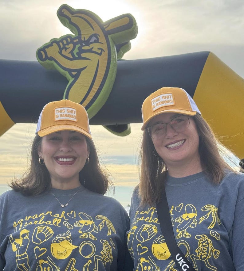 Puede incluir: Dos mujeres con gorras de camionero amarillas y blancas con el texto "THIS SHIT IS BANANAS" y camisetas grises con gr&aacute;ficos de b&eacute;isbol. Un gran arco inflable con forma de pl&aacute;tano en el fondo.