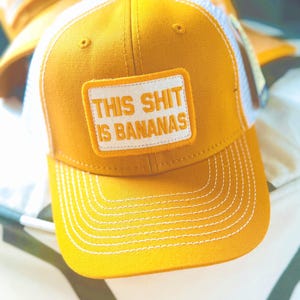 May include: Mustard yellow baseball cap with a white mesh back. The front features a rectangular patch with the phrase "THIS SHIT IS BANANAS" in white block letters. White stitching accents the brim and patch.