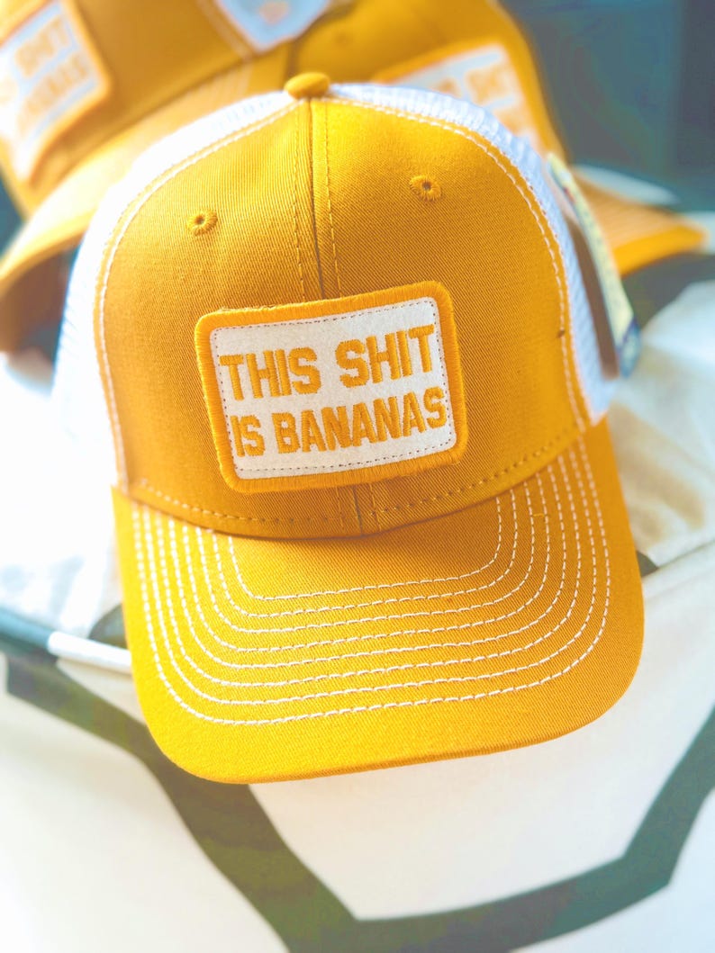 Puede incluir: Gorra de camionero de color amarillo mostaza con la parte trasera de malla blanca y visera curva. La gorra presenta un parche rectangular con la frase "THIS SHIT IS BANANAS" en letras blancas.