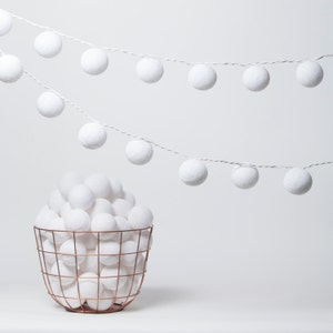 May include: A white wire string of lights with round cotton balls, hanging from the ceiling. A copper wire basket filled with white cotton balls sits on a white surface.
