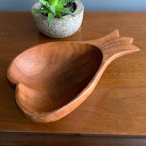 May include: A heart-shaped wooden bowl with a carved handle. The bowl is made of light brown wood and has a smooth finish. The bowl is sitting on a wooden table.