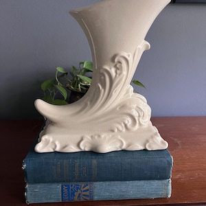 May include: A white ceramic cornucopia vase with a decorative floral design. The vase is sitting on top of a stack of blue books.