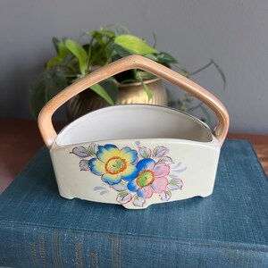 May include: A small, white ceramic planter with a handle. The planter has a floral design with blue, yellow, and pink flowers. The planter is sitting on top of two blue books.