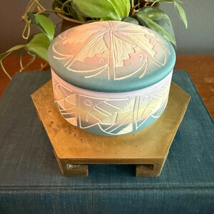 May include: A round, lidded trinket box with a teal and lavender color scheme. The box features an Art Deco design with etched lines and geometric shapes. It sits on a gold-toned hexagonal stand, atop a blue book.