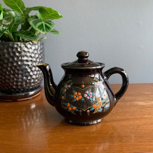 May include: A brown teapot with a floral design in shades of orange, pink, and blue. The teapot has a heart-shaped design in the center with a gold outline.