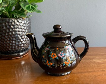 May include: A brown teapot with a floral design in shades of orange, pink, and blue. The teapot has a heart-shaped design in the center with a gold outline.
