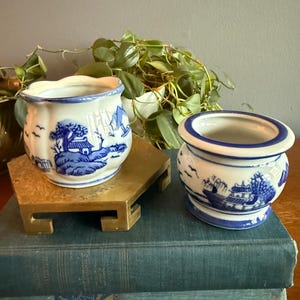 May include: Two small, white and blue ceramic pots with a landscape design. One pot sits on a small, gold-colored stand, and both are placed on top of a stack of books. The pots have a classic, vintage aesthetic.