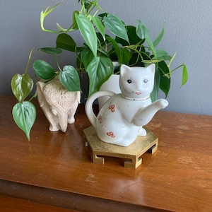 May include: White porcelain teapot shaped like a cat with red floral details, sitting on a gold hexagonal stand.
