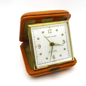 May include: A square travel clock with a gold-tone metal frame and a white face featuring the text "Phinney-Walker", "Eight Days", and "Seven Jewels". The clock is housed in a textured orange leather-like folding case with a hinged lid.