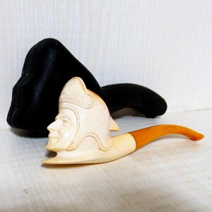 May include: An off-white tobacco pipe with a carved face of a man wearing a helmet. The stem is orange and cream-colored. A black velvet pouch is behind the pipe.