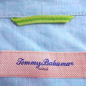 K&ouml;nnte beinhalten: Nahaufnahme eines hellblauen Leinenhemdes mit einem gewebten Etikett mit dem Schriftzug "Tommy Bahama" in blauer Schrift. Ein gr&uuml;n-gelbes Webdetail ist in der N&auml;he des Kragens zu sehen.