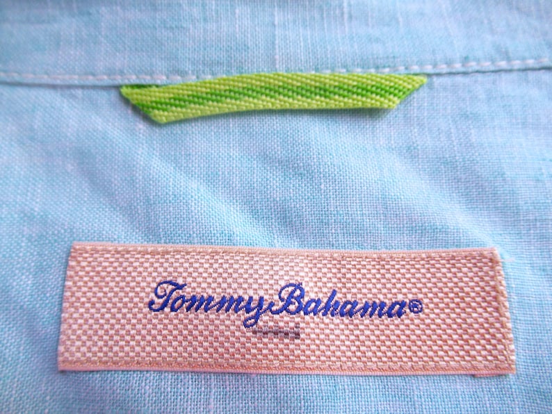 K&ouml;nnte beinhalten: Nahaufnahme eines hellblauen Leinenhemdes mit einem gewebten Etikett mit dem Schriftzug "Tommy Bahama" in blauer Schrift. Ein gr&uuml;n-gelbes Webdetail ist in der N&auml;he des Kragens zu sehen.