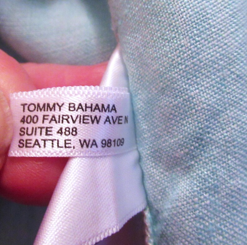 K&ouml;nnte beinhalten: Nahaufnahme eines wei&szlig;en Etiketts mit schwarzem Text: "TOMMY BAHAMA 400 FAIRVIEW AVEN SUITE 488 SEATTLE, WA 98109". Das Etikett ist an einem hellblauen Stoff befestigt.