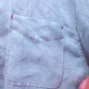 K&ouml;nnte beinhalten: Nahaufnahme eines hellblauen Leinenhemdes mit einer Tasche. Die Tasche ist mit rosa N&auml;hten umrandet, und die oberen Ecken der Tasche haben kleine rote Stiche. Der Stoff des Hemdes hat ein leicht strukturiertes Aussehen.