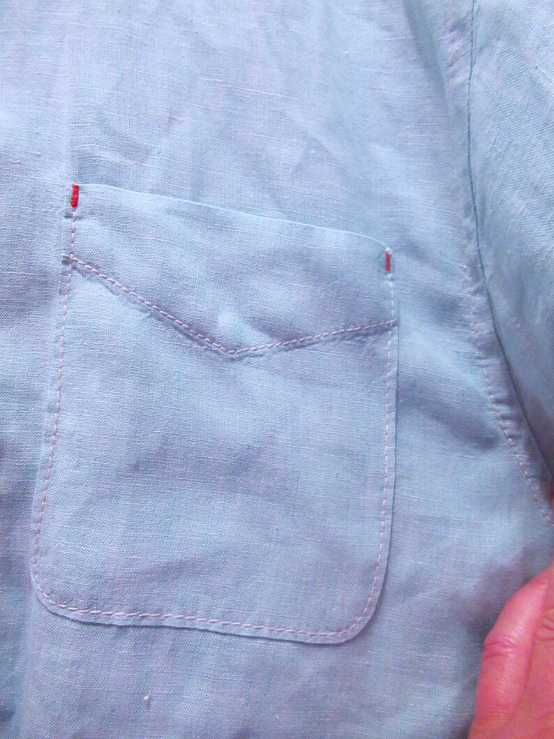 K&ouml;nnte beinhalten: Nahaufnahme eines hellblauen Leinenhemdes mit einer Tasche. Die Tasche ist mit rosa N&auml;hten umrandet, und die oberen Ecken der Tasche haben kleine rote Stiche. Der Stoff des Hemdes hat ein leicht strukturiertes Aussehen.