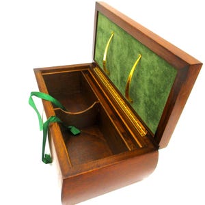 May include: A wooden box with a hinged lid, open to reveal a green velvet interior and gold-colored handles. The box is made of dark wood and has a curved front. A green ribbon is attached inside the box.