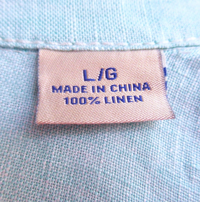 K&ouml;nnte beinhalten: Nahaufnahme eines hellblauen Leinenhemdes. Das Etikett lautet "L/G MADE IN CHINA 100% LINEN". Der Stoff hat ein strukturiertes Aussehen. Das Hemd ist wahrscheinlich ein Freizeitkleidungsst&uuml;ck.