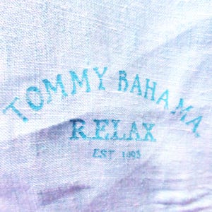 K&ouml;nnte beinhalten: Nahaufnahme eines hellblauen Stoffes mit dem Text "TOMMY BAHAMA RELAX EST 1993" in dunklerem Blau aufgedruckt. Der Stoff hat ein strukturiertes Aussehen.