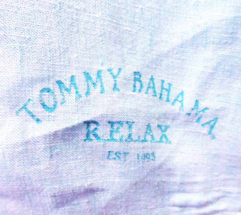 K&ouml;nnte beinhalten: Nahaufnahme eines hellblauen Stoffes mit dem Text "TOMMY BAHAMA RELAX EST 1993" in dunklerem Blau aufgedruckt. Der Stoff hat ein strukturiertes Aussehen.