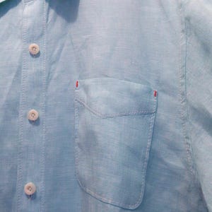 K&ouml;nnte beinhalten: Hellblaues Hemd mit Knopfleiste und Brusttasche. Das Hemd hat wei&szlig;e Kn&ouml;pfe und N&auml;hte, mit roten Akzenten auf der Tasche. Der Stoff scheint Leinen oder ein &auml;hnliches leichtes Material zu sein, das f&uuml;r warmes Wetter geeignet ist.