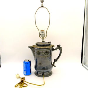 May include: An antique silver-toned lamp with a decorative floral design, featuring a teapot-shaped base and a metal lamp shade frame. The lamp includes a power cord and a blue can for size reference.