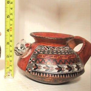 May include: A hand-painted, red ceramic pitcher with a cat-shaped spout and handle. The pitcher features geometric patterns in black, white, and orange, along with insect illustrations. The pitcher measures approximately 5 inches in height.