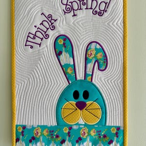 May include: A white quilted wall hanging with a yellow border. The center of the hanging features a blue bunny with purple ears and a yellow nose. The bunny is peeking out from behind a turquoise fabric with a floral pattern. The text "Think Spring!" is embroidered in purple on the top of the hanging.