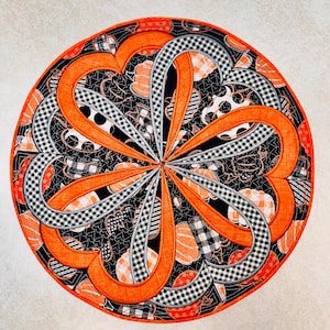 May include: A circular fabric table mat featuring a quilted design with a black background patterned with orange pumpkins. The mat displays a radiating petal motif in solid orange and black-and-white gingham check fabric with orange trim.