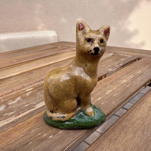 May include: A ceramic figurine of a seated dog with a tan body, red-tipped ears, and black facial markings. The dog sits on a green base. The figurine is placed on a wooden table.
