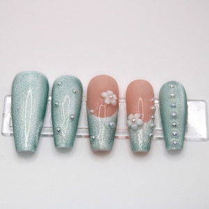 May include: Set of six long, oval-shaped artificial nails. The nails are a light blue color with a shimmery finish. Some nails have white pearl embellishments and white flower accents.