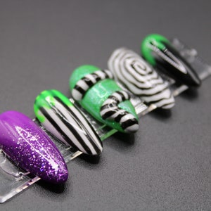 May include: A set of five artificial nails with a variety of designs. One nail is purple with glitter, another has green and white stripes, and a third has a green glitter base with black and white stripes. The other two nails have a black and white swirl and a black and green design.