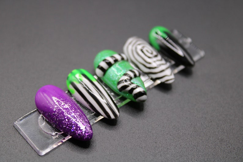 May include: A set of five artificial nails with a variety of designs. One nail is purple with glitter, another has green and white stripes, and a third has a green glitter base with black and white stripes. The other two nails have a black and white swirl and a black and green design.