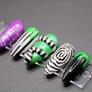 May include: A set of five artificial nails with various designs. One nail is purple glitter, another has black and white stripes, and a third is green with black stripes. The fourth nail has a black and white spiral design, and the last is black with a green tip.
