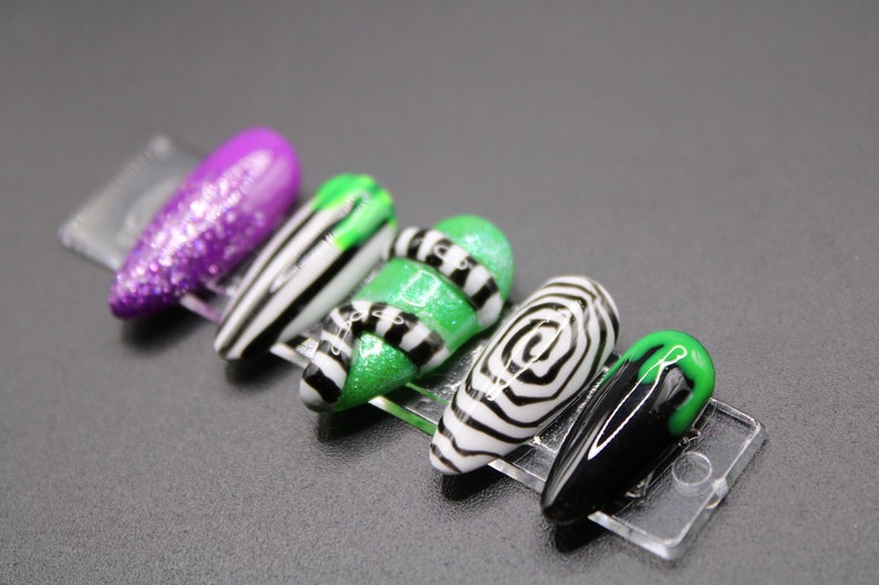 May include: A set of five artificial nails with various designs. One nail is purple glitter, another has black and white stripes, and a third is green with black stripes. The fourth nail has a black and white spiral design, and the last is black with a green tip.