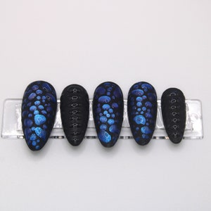 May include: Set of six black press-on nails with a blue and black abstract design. The nails are in a clear plastic holder.