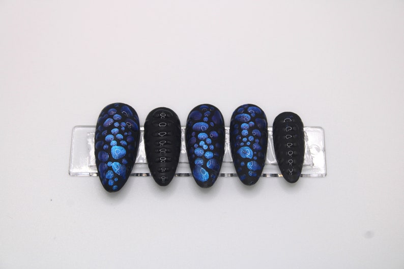 May include: Set of six black press-on nails with a blue and black abstract design. The nails are in a clear plastic holder.