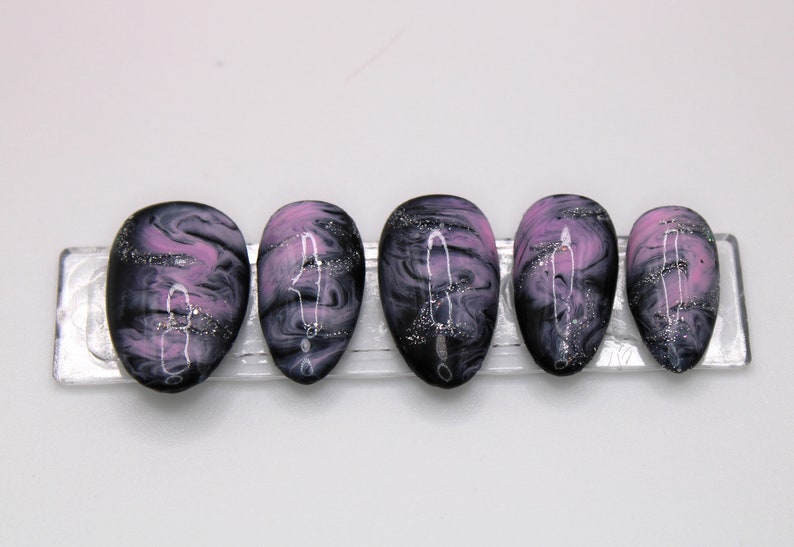 May include: Five black and pink swirl press-on nails with silver glitter. The nails are oval shaped and have a marbled design.
