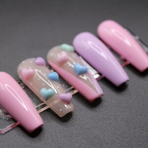 Op de afbeelding: Set van zes lange, roze en paarse kunstnagels met een transparante basis en 3D-hartversieringen in roze, blauw en lichtblauw.