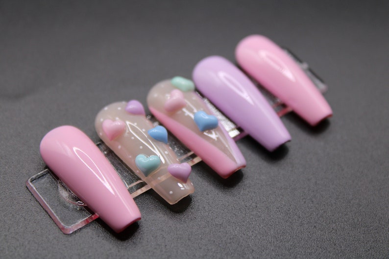 Op de afbeelding: Set van zes lange, roze en paarse kunstnagels met een transparante basis en 3D-hartversieringen in roze, blauw en lichtblauw.