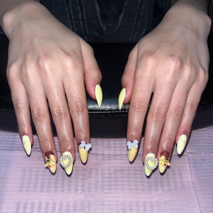 May include: Close-up of hands with yellow almond-shaped nails decorated with white bows, gold hearts, and pearl accents.