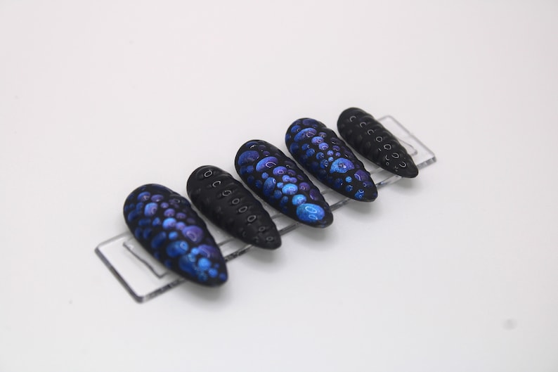 May include: Five black and blue false nails with a textured, dotted design. The nails are arranged on a clear plastic display.