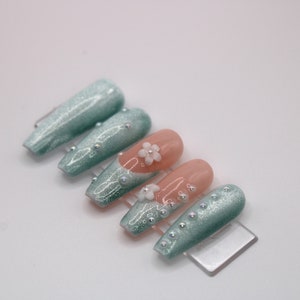 May include: Set of long, light blue and peach-colored press-on nails with glitter and pearl accents. Some nails have a white flower design.