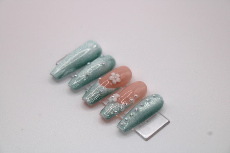 May include: Set of long, light blue and peach-colored press-on nails with glitter and pearl accents. Some nails have a white flower design.