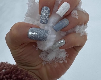 May include: A hand holding snow, showcasing a set of winter-themed nail art. The nails feature a variety of designs, including a snowflake, a cable knit pattern, and glitter accents in shades of gray and white. The nails are long and square-shaped.