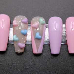 Op de afbeelding: Set van zes lange, roze, lavendel en nude valse nagels met een wit stippenpatroon en 3D hartversieringen in roze, blauw en groen.