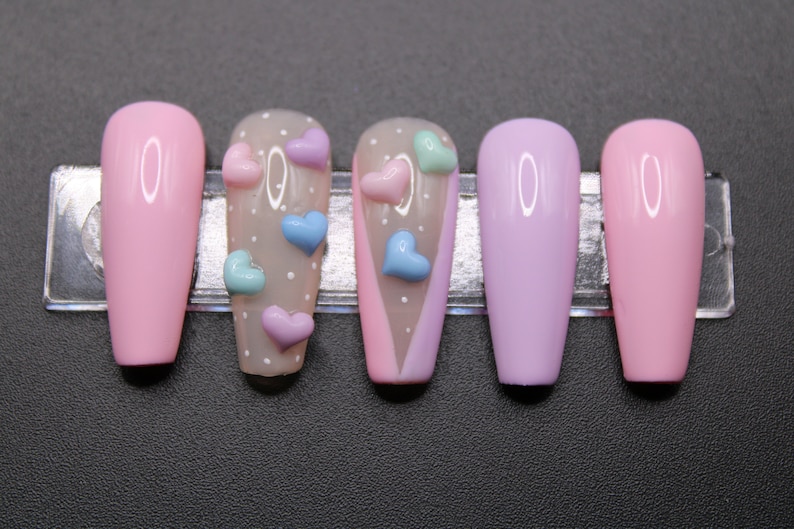 Op de afbeelding: Set van zes lange, roze, lavendel en nude valse nagels met een wit stippenpatroon en 3D hartversieringen in roze, blauw en groen.