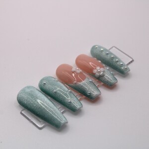 May include: Set of ten press-on nails with a teal glitter finish. Some nails have a white flower accent with pearl embellishments.
